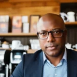 A professional man, identified as author Scott Evans, in a blue suit and glasses sitting thoughtfully in a cafe with shelves and coffee equipment in the background.