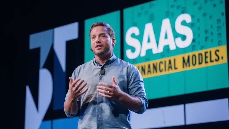 Man presenting on stage at a conference with a slide about "SaaS financial models" displayed in the background. He is using hand gestures while speaking.